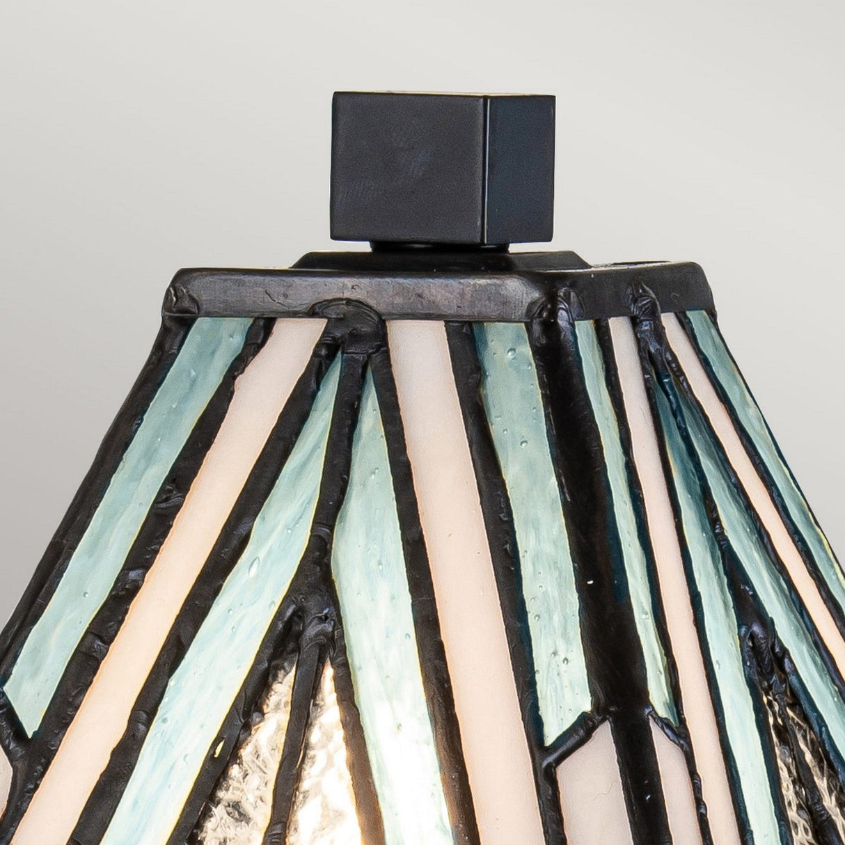 Close-up of the Alderley 1 Light Small Table Lamp, showcasing its Tiffany-style stained glass shade with a geometric design in shades of teal, cream, and gray. This decorative lighting piece features a Vintage Bronze finish and is topped with a black, square-shaped finial.