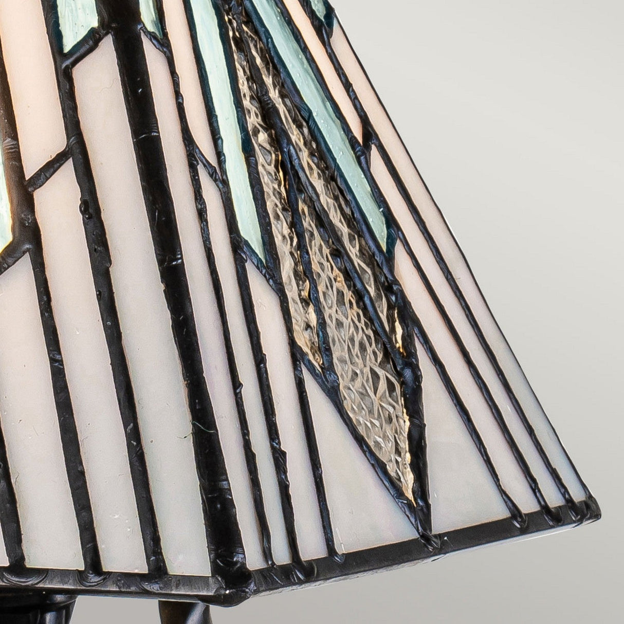 Close-up of the Alderley 1 Light Small Table Lamp's Tiffany-style shade, featuring geometric patterns in shades of white, blue, and black. This decorative lamp includes textured glass sections, lending it an intricate and artistic appearance reminiscent of vintage bronze elegance.