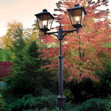 The Turin Large 2 Light Twin Lamp Post - Black, made from robust die-cast aluminum, stands gracefully amidst vibrant autumn trees adorned with red, orange, and green leaves. This picturesque scene is beautifully highlighted against a red fence under a soft evening sky, exemplifying sophisticated outdoor illumination.