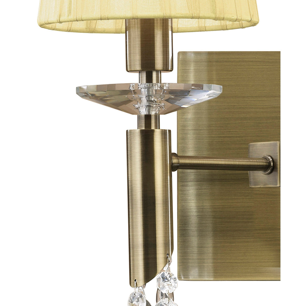 Close-up of Tiffany wall light detail showing antique brass arm, faceted crystal disc, and cream pleated shade against a plain white background.
