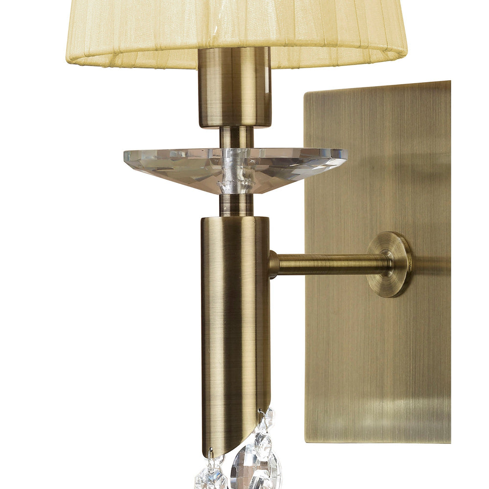 Close-up of Tiffany wall light showing antique brass arm, rectangular backplate, cream pleated shade and faceted crystal detail on a white background.