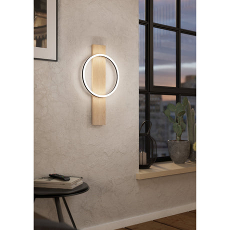 Timarne LED wall light in black and brown, with a round ring crossing a tall oak-toned wood backplate, mounted beside a window in a living room setting.