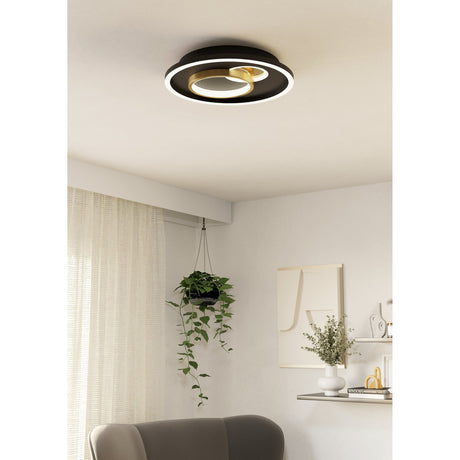 Tiriolo LED flush ceiling light in black and gold, round steel and plastic disc with white outer diffuser and overlapping gold rings, shown mounted on a living room ceiling above a hanging plant.