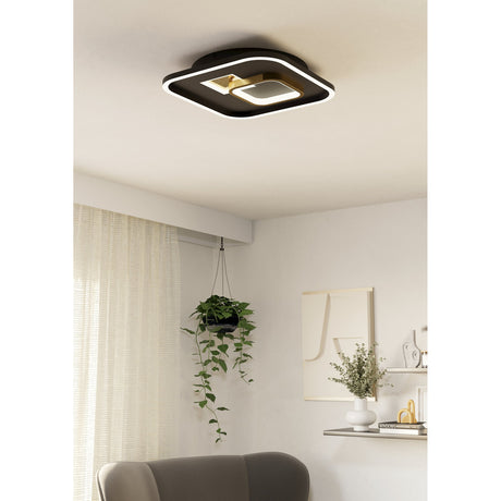Tiriolo square LED ceiling panel light in black and gold, with rounded corners, white glowing edge and gold crackle-effect insert, shown mounted in a living room with sheer curtains and hanging plant.
