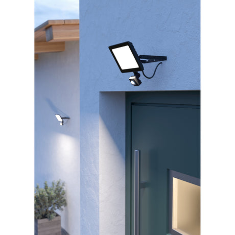 Tirnine LED flood light with motion sensor in black aluminium, wall-mounted beside a front door, with a second unit visible illuminating the house exterior in the background.