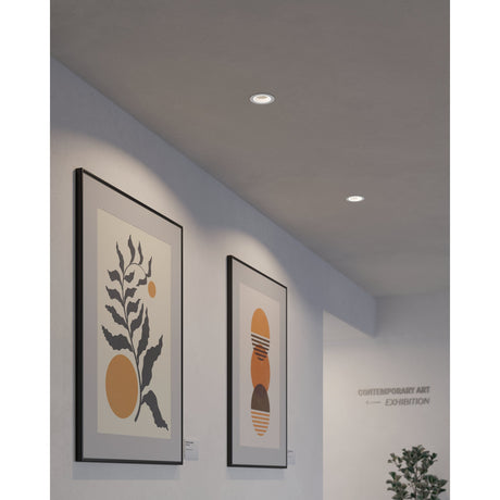 Two round Tomelle 9.3cm LED downlights recessed into a white ceiling, with white bezels, shown above framed artwork in a gallery-style corridor.