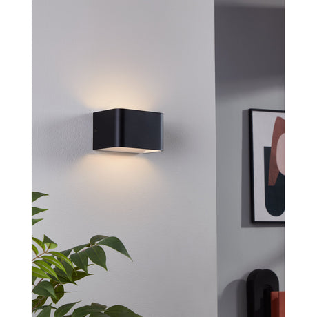 Tomessa LED Wall Light in matt black aluminium with rounded edges, mounted on a grey wall, casting warm white uplight and downlight beside a framed print and plant.