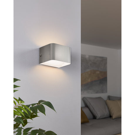 Tomessa LED Wall Light 13cm in satin nickel aluminium, a compact rectangular up-and-down wall fixture with white interior, shown illuminated on a light grey wall with living room setting visible.