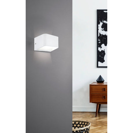 Tomessa LED Wall Light 13cm in white aluminium with a rounded cube shape, mounted on a grey wall beside a living space with artwork and a wooden sideboard.