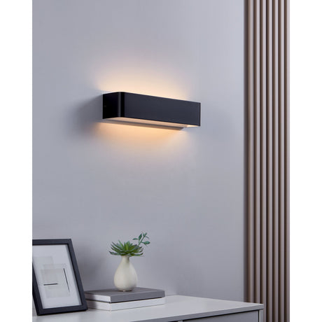 Tomessa LED Wall Light in black aluminium with white diffuser, 36.5cm rectangular design casting warm uplight and downlight on a bedroom wall above a console table.