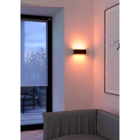 Tomessa black rectangular LED wall light casting warm upward and downward glow beside a patio door in a living room with grey sofa and framed art.