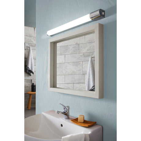 Tragacete 1 LED bathroom wall light mounted above a framed mirror, showing its slim white tube and chrome end caps against a pale blue bathroom wall.