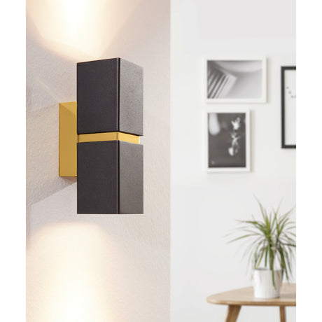 Tramena Wall Light 5.5cm in black and gold, featuring two square steel blocks on a gold backplate with up and down lighting, shown mounted on a wall beside framed artwork.