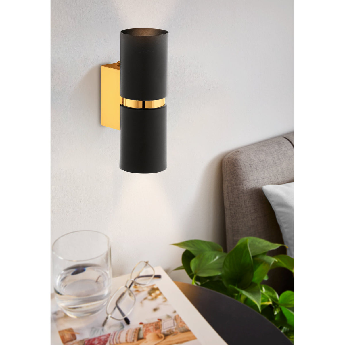 Tramena Wall Light 6cm in black and gold, two matt black steel cylinders joined by a polished gold band on a gold backplate, mounted above a bedside table in a bedroom setting.