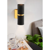 Tramena Wall Light 6cm in black and gold, two matt black steel cylinders joined by a polished gold band on a gold backplate, mounted above a bedside table in a bedroom setting.