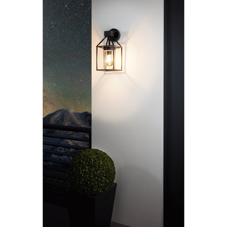 Trecate Outdoor Wall Light in black, a lantern-style fixture with a square steel frame and clear glass cylinder, shown illuminated on an exterior wall beside a balcony and potted plant at night.