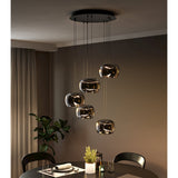 Hortunas 5-light cluster pendant with round black steel ceiling canopy and smoked glass shades staggered on varied cables, shown above a dining table in a dark-toned room.