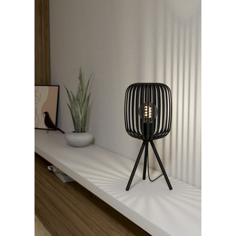 Trosvelle Table Lamp in black steel with a slatted cage shade and tripod legs, shown lit on a white shelf beside a plant and framed art.