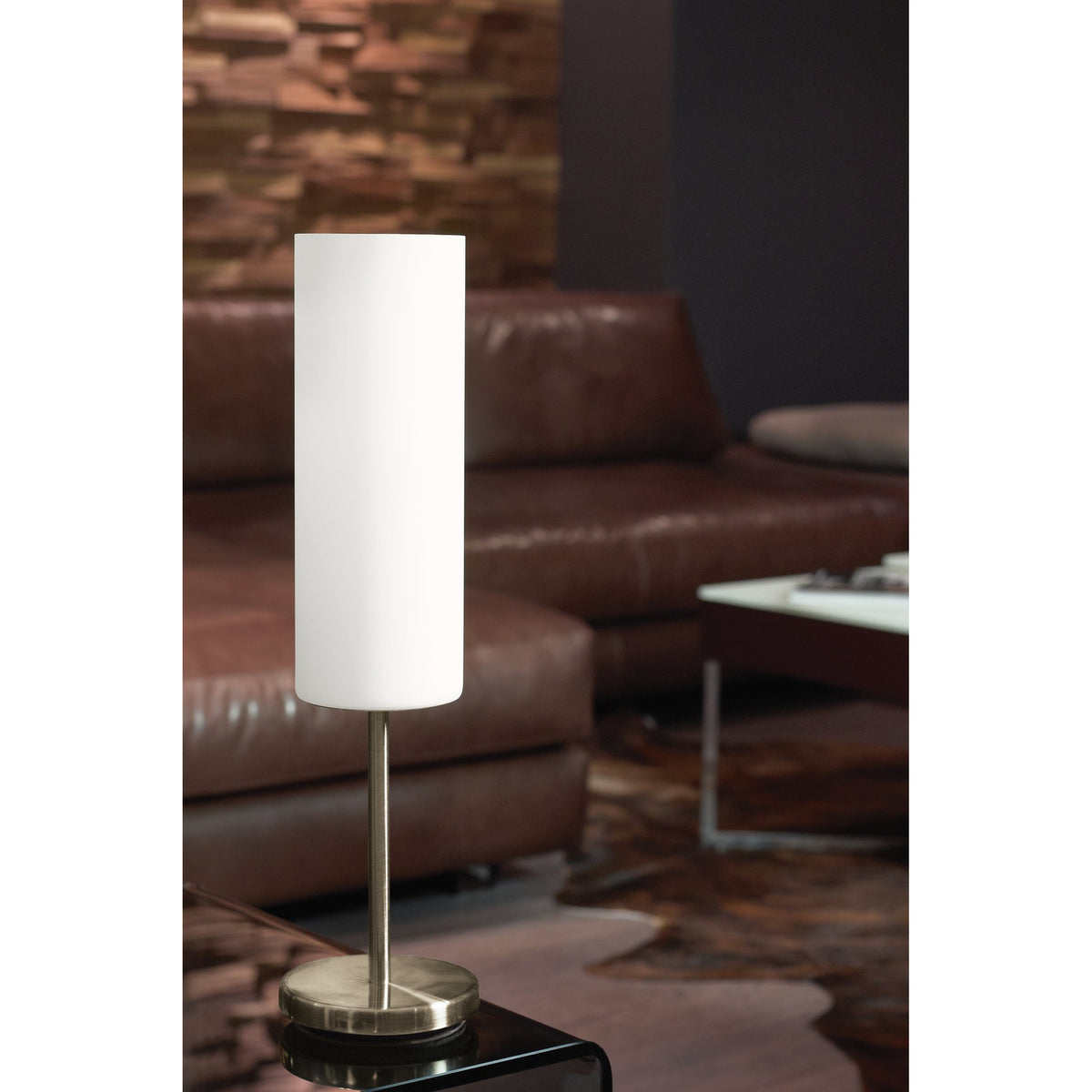 Troy 3 Table Lamp in satin nickel with a tall cylindrical white glass shade, shown on a side table beside a brown leather sofa in a living room setting.