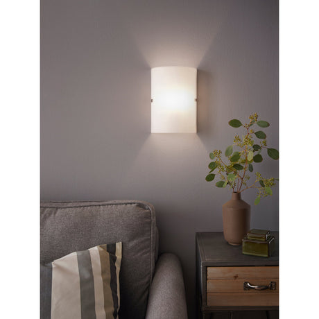 Troy 3 Wall Light in Satin Nickel with a curved frosted glass panel, illuminated on a grey living room wall above a side table with a vase and plant.
