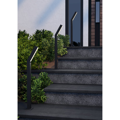 Ugento LED bollard lights in matte black with angled heads and white diffusers, shown lining stone steps beside greenery at a house entrance.