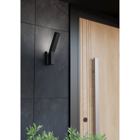 Ugento LED Outdoor Wall Light in black, a slim angled steel bar fixture mounted on a dark tiled wall beside a timber front door, casting a soft downward glow.