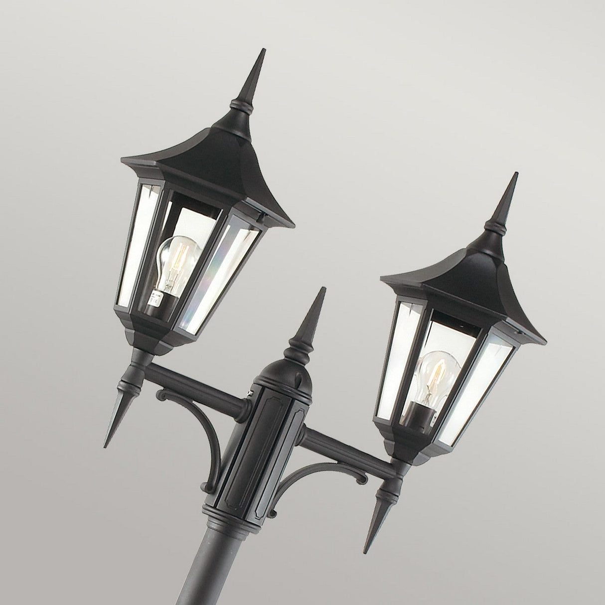 The Valencia 2 Light Twin Head Lamp Post in black, featuring pointed tops and clear glass panes, beautifully illuminates garden pathways. The visible light bulbs offer elegant outdoor lighting against a plain gray backdrop.