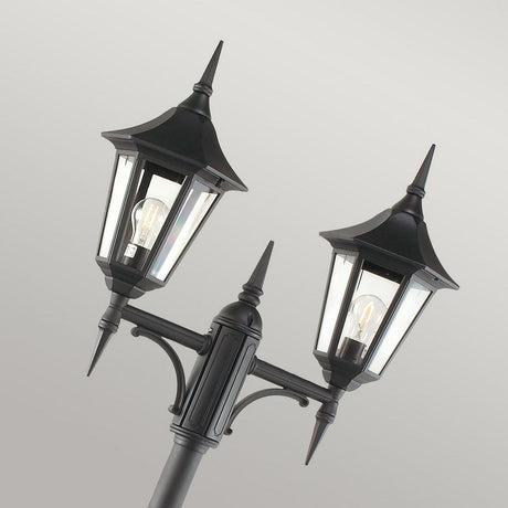 The Valencia 2 Light Twin Head Lamp Post in black, featuring pointed tops and clear glass panes, beautifully illuminates garden pathways. The visible light bulbs offer elegant outdoor lighting against a plain gray backdrop.