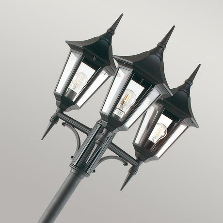 The Valencia 3 Light Triple Head Lamp Post - Black is an intricate outdoor lighting fixture featuring clear glass panels and visible bulbs, elegantly contrasted against a simple, light gray background.