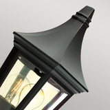 A close-up of the Valencia Outdoor 1 Light Half Lantern - Black showcases its sleek black design, reminiscent of vintage outdoor lighting. The clear glass panels reveal the visible bulb inside, while the lamp's pointed top adds a touch of sophistication against a neutral backdrop.