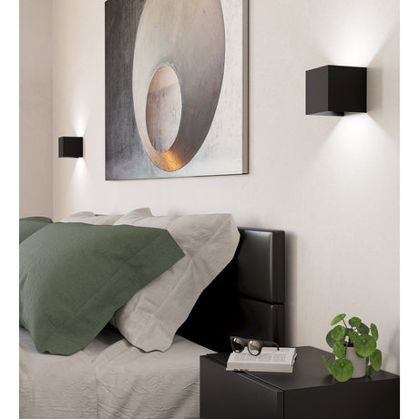 Valmontone LED Wall Light in black aluminium, cube-shaped fixtures mounted either side of a bed, casting warm white light up and down the wall in a bedroom setting.