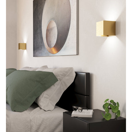 Valmontone LED Wall Light in brushed brass, cube-shaped aluminium fixture mounted above a bedside nightstand, casting light up and down the wall beside framed artwork.