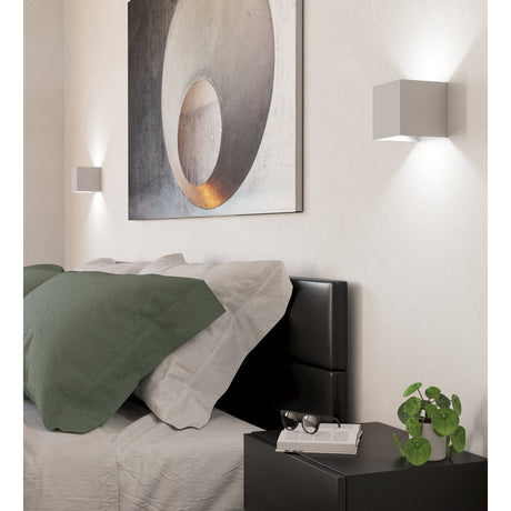 Valmontone LED Wall Light in white aluminium cube design, shown on bedroom walls with up-down lighting above a bed and bedside table.