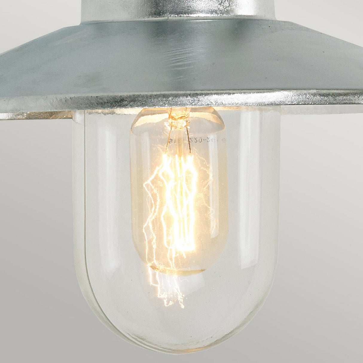 A close-up of the Vansbro Outdoor 1 Light Wall Lantern in silver shows its illuminated bulb encased in an IP55 rated galvanized steel frame. The glowing filament inside emits a warm light, beautifully contrasting with the metallic top. A simple, light gray background enhances the lantern's elegant design.