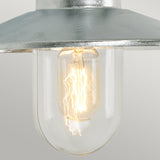 A close-up of the Vansbro Outdoor 1 Light Wall Lantern in silver shows its illuminated bulb encased in an IP55 rated galvanized steel frame. The glowing filament inside emits a warm light, beautifully contrasting with the metallic top. A simple, light gray background enhances the lantern's elegant design.