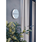 Vascella LED Wall Light 18cm in white stainless steel, round split-disc design glowing softly on a grey exterior wall beside a curtained glass door.
