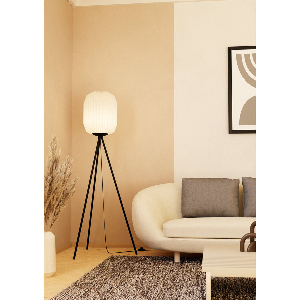 Vavanne tripod floor lamp with black steel legs and a tall pleated white paper lantern shade, shown lit beside a cream sofa in a living room.