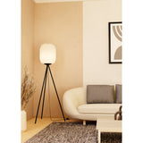 Vavanne tripod floor lamp with black steel legs and a tall pleated white paper lantern shade, shown lit beside a cream sofa in a living room.