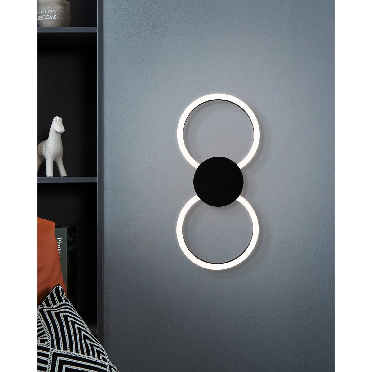 Venticello 1 LED Flush Ceiling Light in black, shown wall-mounted as two illuminated rings joined by a round disc, used as a hallway wall light beside a dark shelving unit.
