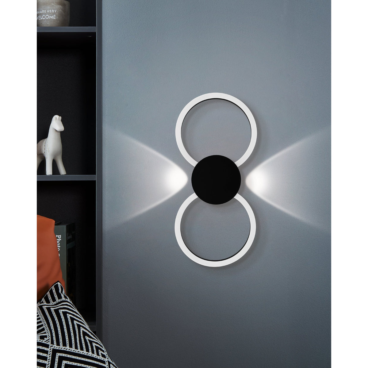 Venticello LED wall light in black, two linked rings with a round central disc, mounted on a grey wall casting soft upward and downward light beams.
