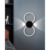 Venticello LED wall light in black, two linked rings with a round central disc, mounted on a grey wall casting soft upward and downward light beams.