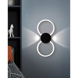 Venticello LED wall light in black, two linked rings with a central disc and glowing white diffusers, mounted on a grey wall above a dark bookshelf.
