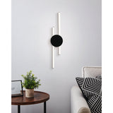 Venticello LED Flush Ceiling Light in black, shown on a living room wall with two offset vertical light bars and a round disc canopy, styled near a sofa and side table.