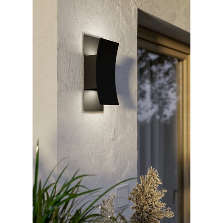 Verdolino LED outdoor wall light in black steel with a curved front panel over a rectangular backplate, mounted beside a door and casting up-and-down light on a textured exterior wall.