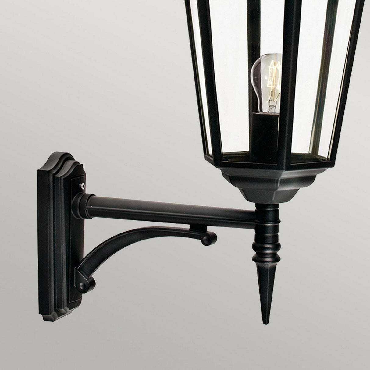 The Valencia Outdoor Large 1 Light Up Lantern - Black is a die-cast aluminum, wall-mounted fixture with a transparent glass enclosure that reveals the visible light bulb inside. Featuring an IP54 rating, its decorative arm elegantly stands out against a plain gray background.
