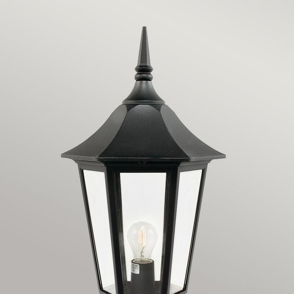 The Valencia Large 1 Light Pedestal - Black is a traditional-style outdoor lantern made from die-cast aluminum. It features a pointed top and showcases a clear glass enclosure that reveals the bulb inside, making it an excellent choice for outdoor lighting. The backdrop is a simple light gray.