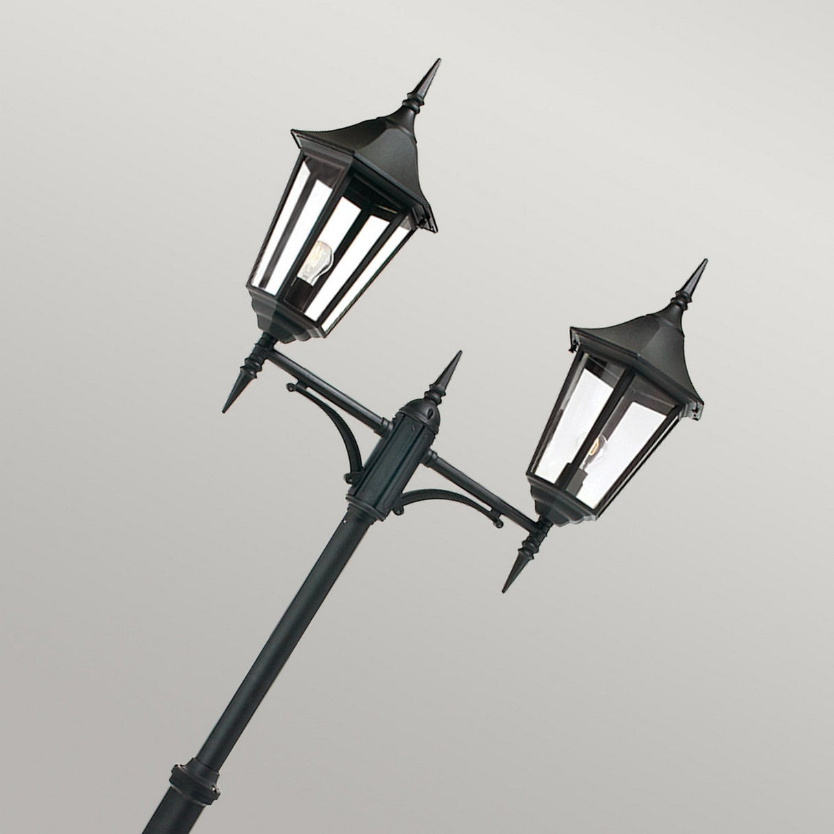 The Valencia Large 2 Light Twin Head Lamp Post - Black features lantern-style lights and stands against a light gray background. Made from die-cast aluminum, it showcases a classic design with pointed finials and clear glass panels, each light displaying a visible bulb. Ideal for outdoor lighting, it radiates timeless elegance.