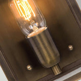 A close-up view of a vintage-style light bulb glowing inside a brushed metal fixture, similar to the Victoria 1 Light Outdoor Wall Light in aged brass, mounted against a dark wooden background. The bulb's filament is clearly visible, casting a warm, ambient glow, and the fixture is IP44 rated for outdoor use.