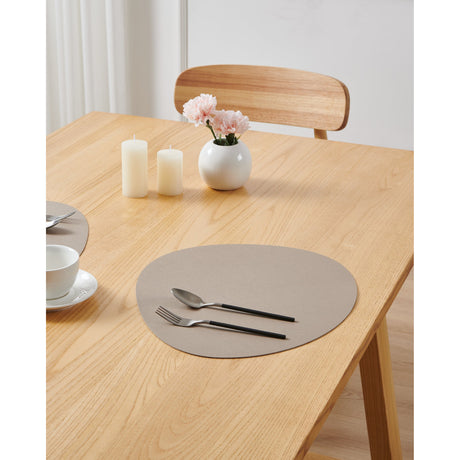 Vinanivao oval placemat in beige, fine-grain plastic finish, styled on a wooden dining table with cutlery, candles, and a flower vase in a bright room.
