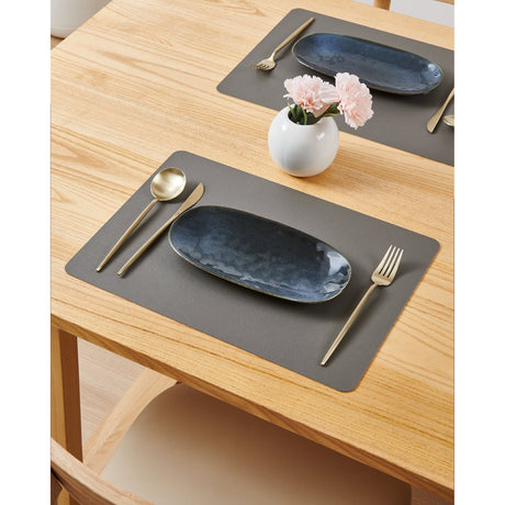 Vinanivao rectangular placemat in dark grey with pebbled, leather-like texture, shown set on a wooden dining table with a blue oval plate, gold cutlery, and a vase of pink flowers.